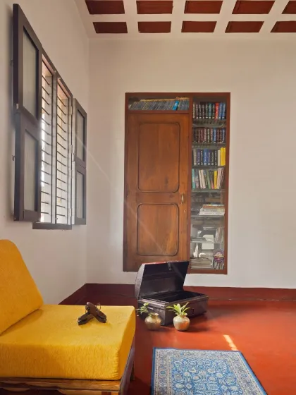 A quiet reading corner in the Mattur residence, featuring an antique book cabinet, a traditional chest, and a simple yellow divan. The louvered window filters soft light onto the earthy terracotta floor.