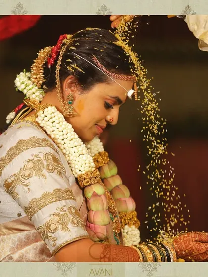 A bride during her wedding ceremony, wearing a custom white blouse with gold lotus embroidery on the sleeves. The design complements her traditional lotus garland.