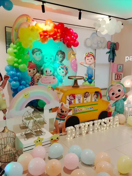A vibrant Cocomelon themed party for a second birthday. The setup is centered around the iconic yellow school bus and features a rainbow balloon arch, character cutouts, and fluffy clouds to recreate the show's happy world.