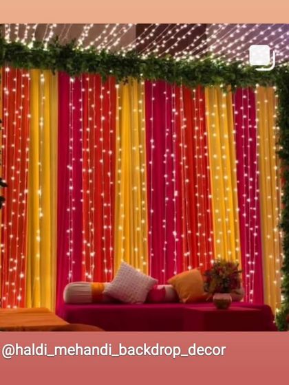A cozy seating area with a backdrop of colorful drapes and fairy lights. The pink and yellow fabric creates a warm and inviting atmosphere for a Sangeet or Mehandi night.