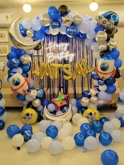An elaborate space-themed birthday party with a full balloon ring in blue, white, and silver. The setup includes a neon sign, foil balloons of the moon, rockets, and an astronaut.
