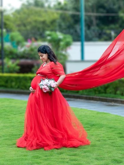 A beautiful outdoor shot of a mom-to-be in a red gown with a long, flowing train, holding a bouquet of flowers.