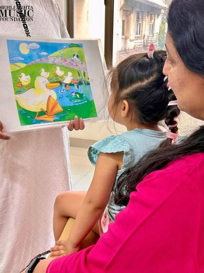 A mother and child look at a colorful picture book during a music class. We use storytelling and visual aids to bring songs and musical concepts to life.