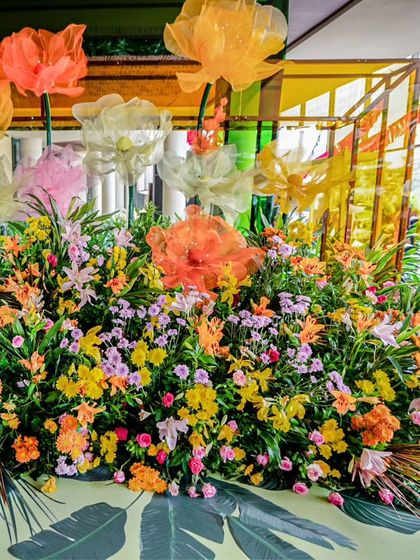A large floral installation at the Haldi, combining fresh, colorful blooms with the oversized fabric flowers, all set against a backdrop of tropical leaf prints.