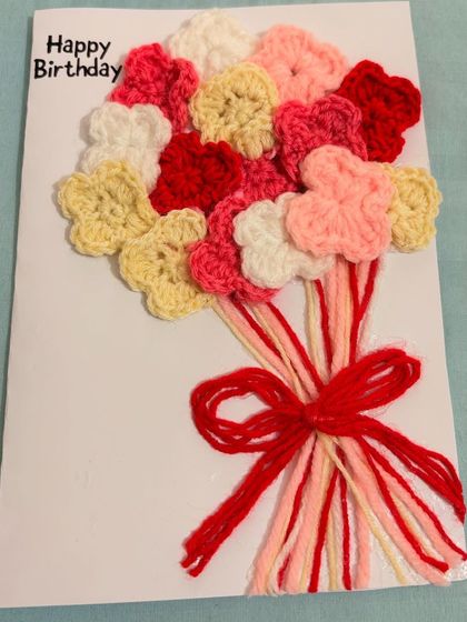 A special birthday card featuring a multi-colored bouquet of crochet flowers tied with a red yarn bow. It's a warm and creative way to send birthday wishes.