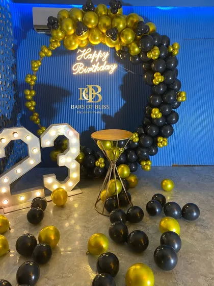 A bold and energetic 23rd birthday celebration. The decor features a full ring of black and yellow balloons against a vibrant blue textured wall, complete with marquee numbers and a neon sign.