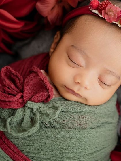 A close-up portrait focusing on the serene expression of this baby girl, surrounded by a bed of red flowers. The floral headband adds the perfect finishing touch.