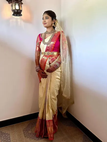 A full-length view of a bride in a stunning cream and pink Kanchipuram saree. Her bright pink blouse is heavily embroidered, creating a beautiful contrast for her muhurtham look.