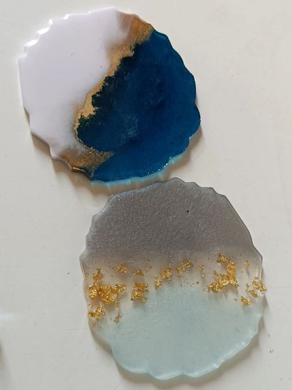A pair of geode-style resin coasters showing two different color schemes. One features a deep blue and white with gold accents, while the other has a soft, shimmery grey with gold flakes.