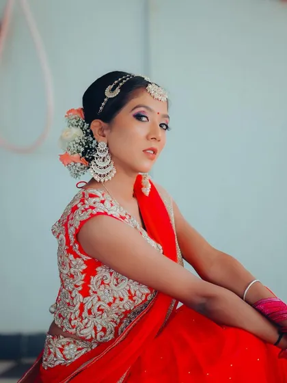 A close-up portrait of a model in a red lehenga, showcasing her elegant hairstyle with flowers and detailed makeup. The soft lighting enhances the romantic feel of the shot.