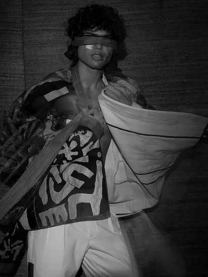 A high-contrast, black and white image from the same artistic campaign. The model is blindfolded, and the focus is on the chaotic, layered patterns of the different fabrics, creating a powerful, moody shot.
