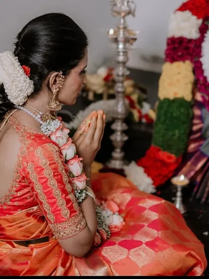 A bride during a pooja ceremony, wearing a beautiful orange and pink saree. The back of her blouse shows delicate embroidery work that complements the saree perfectly.