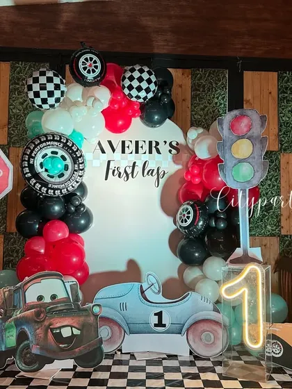 We brought the excitement of the racetrack to this 'First Lap' birthday celebration. The decor includes a custom arch backdrop, balloon garlands in red, black, and white, checkered flag details, tire props, and character cutouts from the movie 'Cars'.