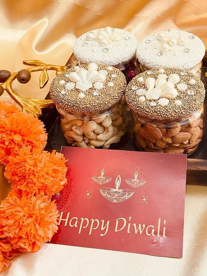 Our decorative dry fruit jars are a festive favorite. This set features four jars with intricately designed lids, filled with premium cashews and almonds, presented on a wooden tray.