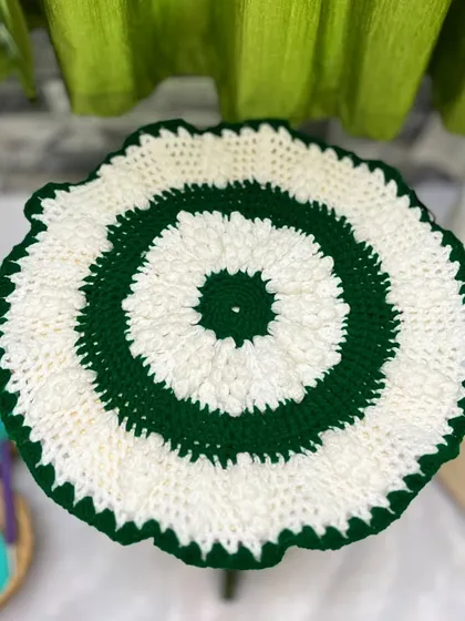 This is a simpler version of a crochet doily or 'rumal', with a puffy white stitch and a green border, sitting on a small table.