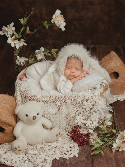 A cozy and sweet newborn setup with a white bunny theme. The baby, wearing a fuzzy bonnet, sleeps peacefully with a teddy bear and floral accents.