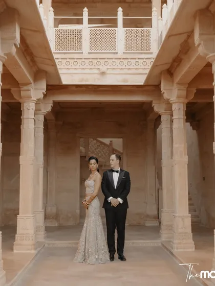 A formal portrait set within the grand, pillared hall of Suryagarh. The symmetrical composition and the couple's poised stance create an image of timeless elegance and sophistication.