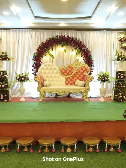 A stage prepared for a traditional function, featuring a circular arch of red roses against a white curtain. The ornate two-toned sofa is the centerpiece, ready for the main event.