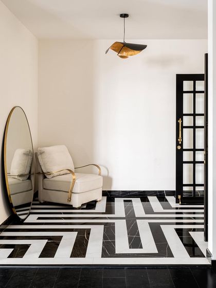 A striking entryway featuring geometric black and white marble flooring. A plush armchair and a large, round brass mirror create an elegant waiting spot, demonstrating how we use bold patterns and reflective surfaces to make a strong first impression.