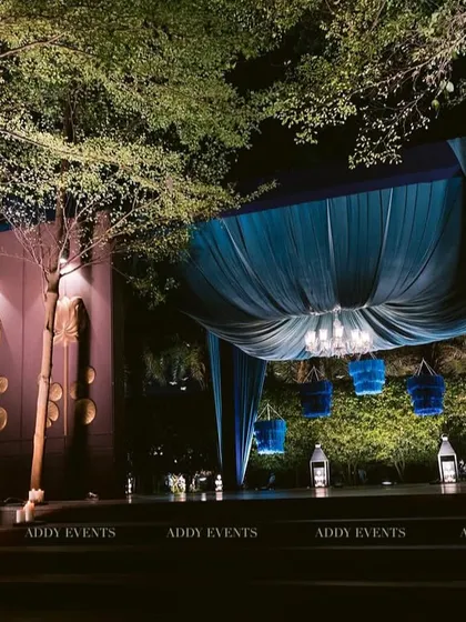 A tranquil and sophisticated stage setting by a grove of trees. The deep blue drapes, hanging lanterns, and surrounding candles transform this outdoor space into an intimate performance area.