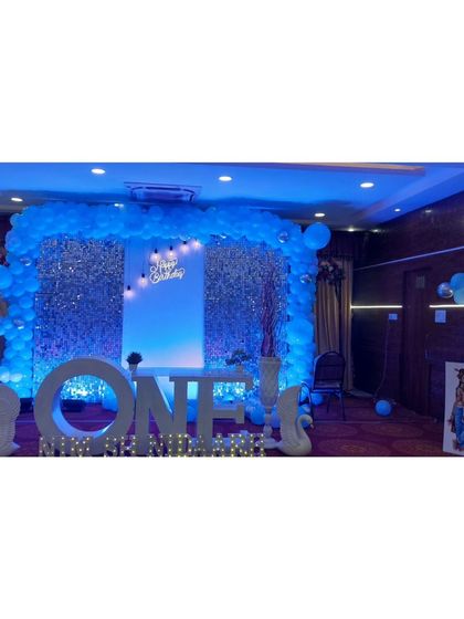 A stunning setup for a first birthday, featuring a large light-up 'ONE' sign as the centerpiece. The backdrop is a silver shimmer wall framed by a blue balloon arch, creating a cool, celebratory glow for the big milestone.