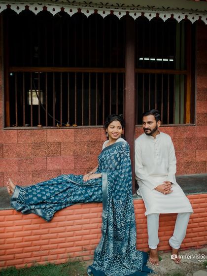 A relaxed and stylish shot of a couple at a traditional house. The bride's beautiful blue printed saree drapes elegantly as she sits on a brick ledge, creating a rustic and charming scene.