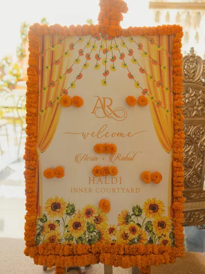 A personalized welcome sign for the Haldi ceremony, beautifully decorated with a border of fresh marigolds and a sunflower motif, directing guests to the inner courtyard.