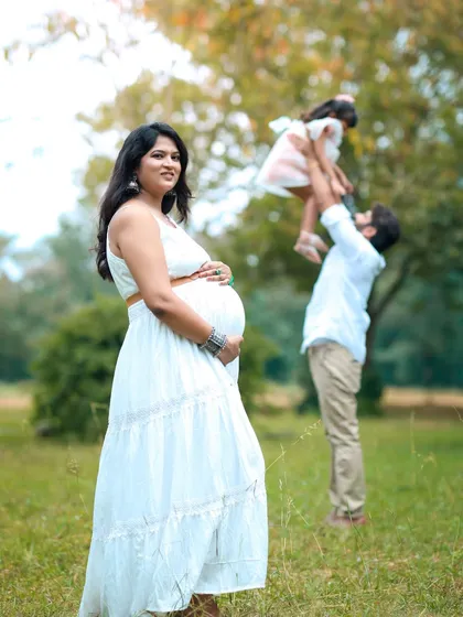 A beautiful outdoor maternity session capturing a candid family moment. The expectant mother is in focus, while in the background, the father joyfully lifts their older child in a sunlit park, telling a story of a growing family.