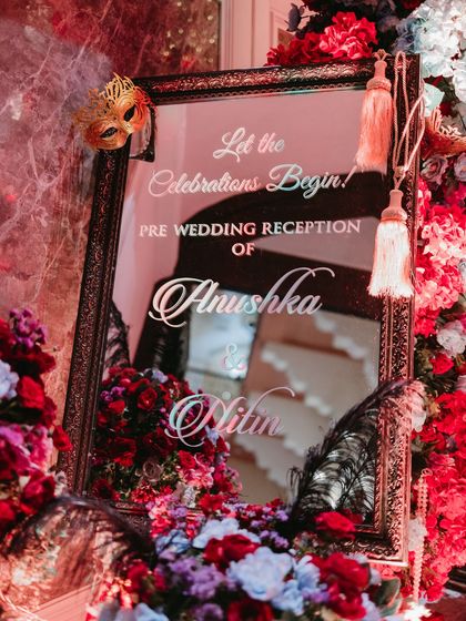 A personalized welcome mirror for the pre-wedding reception, framed with lush flowers and a decorative mask, inviting guests into the celebration.