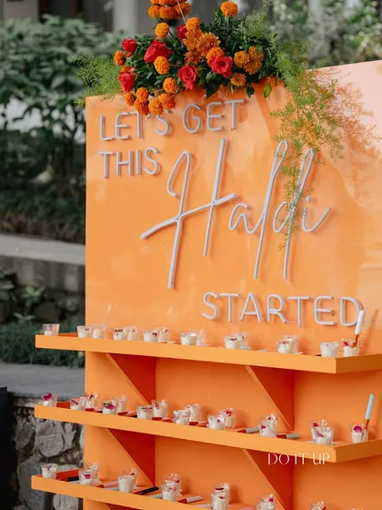 An interactive and playful element for a Haldi by the Ganges. This custom orange stand, reading "Let's Get This Haldi Started," holds individual portions of ubtan for guests.