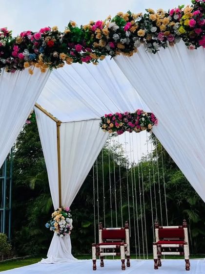 A simple and elegant outdoor wedding mandap with a white theme. The structure is draped with white fabric and adorned with colorful floral bouquets, perfect for a day wedding.