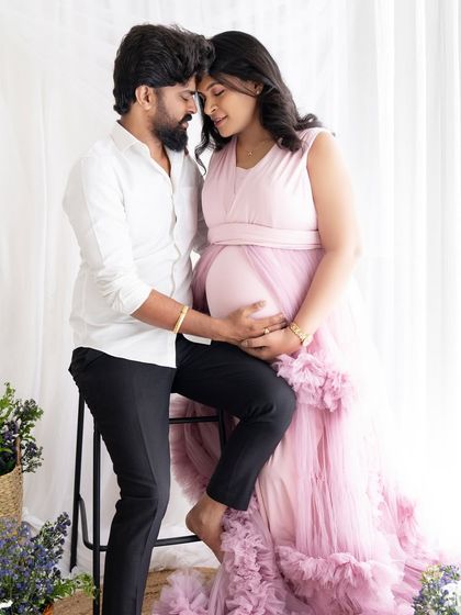 A romantic pose with the couple close together, the mom-to-be seated while her partner stands beside her, both dressed in a soft pink and white palette.