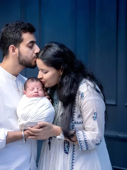 A classic pose for a new family. The parents' gentle kisses on the baby's head create a beautiful silhouette against the dark studio background, highlighting their affection.