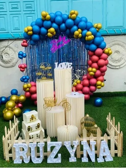 A vibrant outdoor birthday setup for Ruzyna. The decoration features a colorful balloon ring against a blue fringe backdrop, a picket fence, and various props on pedestals.