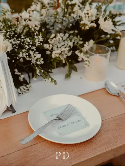 A detail of the place setting, with a personalized card and a simple white plate. The focus is on clean elegance and the beauty of the surrounding nature.