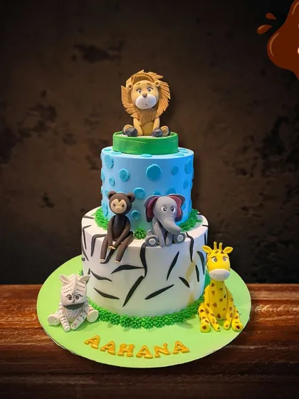 This two-tier safari cake features a zebra-print bottom layer and a blue-dotted top layer. It's adorned with a variety of hand-molded fondant animals, including a lion, monkey, elephant, and giraffe.
