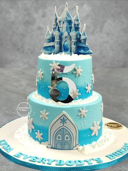A magnificent two-tiered Frozen castle cake. This design features a detailed, edible ice castle on top, with snowflake decorations all around.