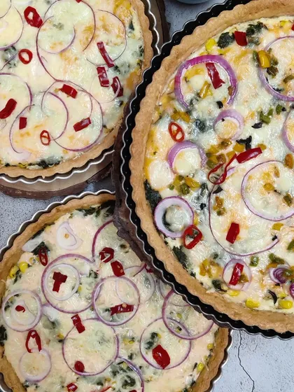 An overhead view of our freshly baked vegetarian quiches, showcasing the flaky crust and generous fillings like spinach, corn, and cheese, topped with red onion and chili.