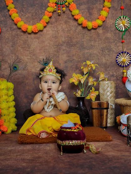 A wide shot of our elaborate Krishna setup, featuring a baby boy enjoying the festive scene. We take pride in creating immersive and beautiful backdrops.