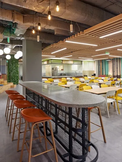 A cafeteria with an industrial vibe, featuring an exposed ceiling, concrete-topped high table, and a mix of seating types. The green wall in the background adds a touch of nature.