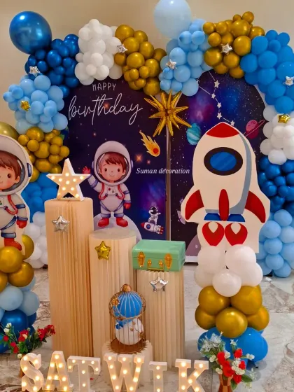 A detailed astronaut and space themed birthday party. The setup includes a galaxy backdrop, cute astronaut cutouts, wooden pedestals for the cake, and a balloon garland in blue, gold, and white.