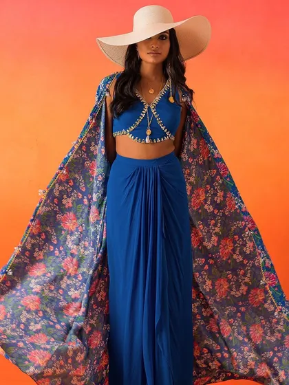 A perfect destination wedding look. This ensemble features a royal blue draped skirt and bralette, paired with a dramatic floral-printed cape and a wide-brimmed hat.