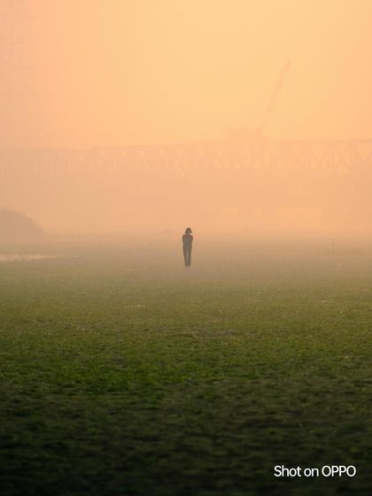 A lone figure stands in a vast, grassy field, enveloped in the thick morning fog with a bridge faint in the background. The image conveys a sense of solitude and mystery.
