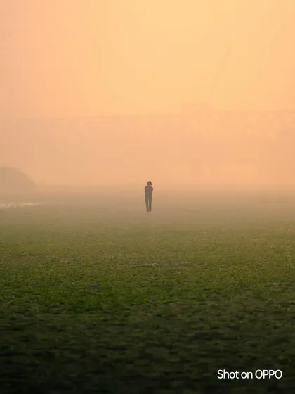 A lone figure stands in a vast, grassy field, enveloped in the thick morning fog with a bridge faint in the background. The image conveys a sense of solitude and mystery.