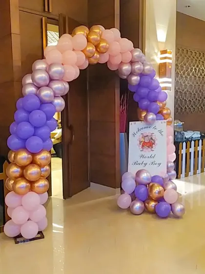 A balloon arch in pastel pink, purple, and gold at the entrance of a "Welcome to the World Baby Boy" party.