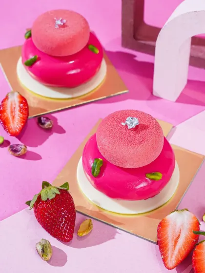 Two glossy pink dome desserts, topped with smaller mousse spheres and garnished with pistachios. The sharp, bright lighting and pink background give this shot a modern, high-end patisserie feel.