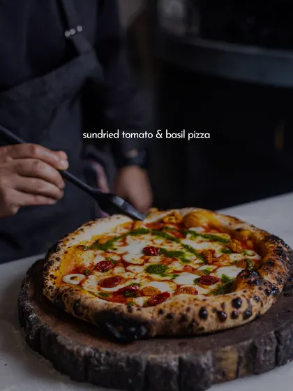 This is what it's all about. A beautiful Sundried Tomato & Basil pizza, fresh from the wood-fired oven. You can see the perfectly charred crust, the melted mozzarella, and the vibrant pesto, all crafted by our pizzaiolo right before your eyes.