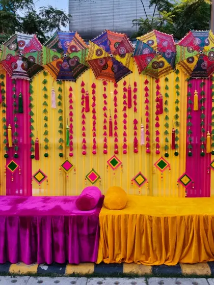 A vibrant stage seating for a Mehendi or Haldi event. The backdrop is a mix of yellow and pink drapes, decorated with colorful umbrellas and traditional tassels.