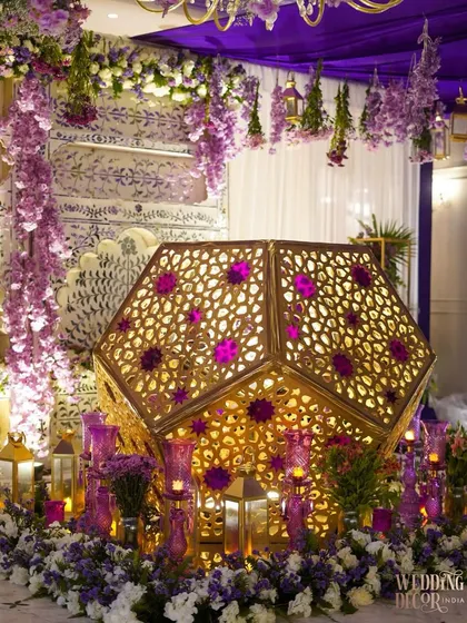 A stunning installation for an evening party, featuring a large, illuminated geometric shape surrounded by a sea of purple and white flowers and candles.