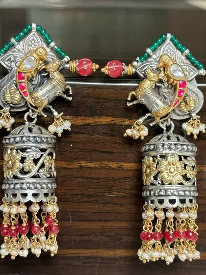 These dual-tone jhumkas are a perfect example of fusion design. They feature intricate silver work with gold-plated accents, colourful stones, and delicate pearl and ruby bead drops.